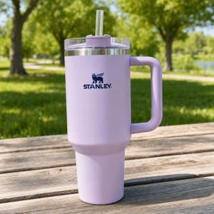 Puede incluir: Un vaso Stanley lavanda con asa, tapa transparente y pajita. El logotipo de Stanley est&aacute; impreso en azul marino. El vaso est&aacute; sobre una superficie de madera con un fondo verde.