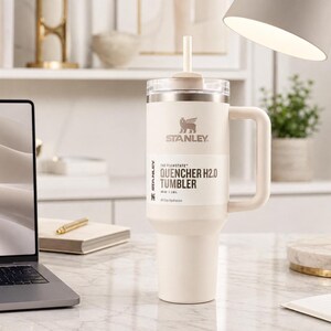 May include: Cream-coloured Stanley Quencher H2.0 Flowstate tumbler with a handle and straw. The tumbler has the Stanley logo and product information printed on the side. It sits on a white marble surface next to a laptop and a desk lamp.
