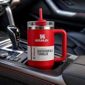 K&ouml;nnte beinhalten: Ein roter Stanley Quencher H2.0 Becher mit Henkel und Strohhalm, ca. 1,18L Fassungsverm&ouml;gen. Der Becher hat das Stanley-Logo und Produktinformationen auf einem wei&szlig;en Etikett. Er steht in einem Getr&auml;nkehalter im Auto.