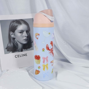 May include: A light blue insulated water bottle with a pink lid and a beige band. The bottle is decorated with illustrations of food and drinks, including a lobster, shrimp, and a martini glass. A black and white photo with the word "CELINE" is in the background.
