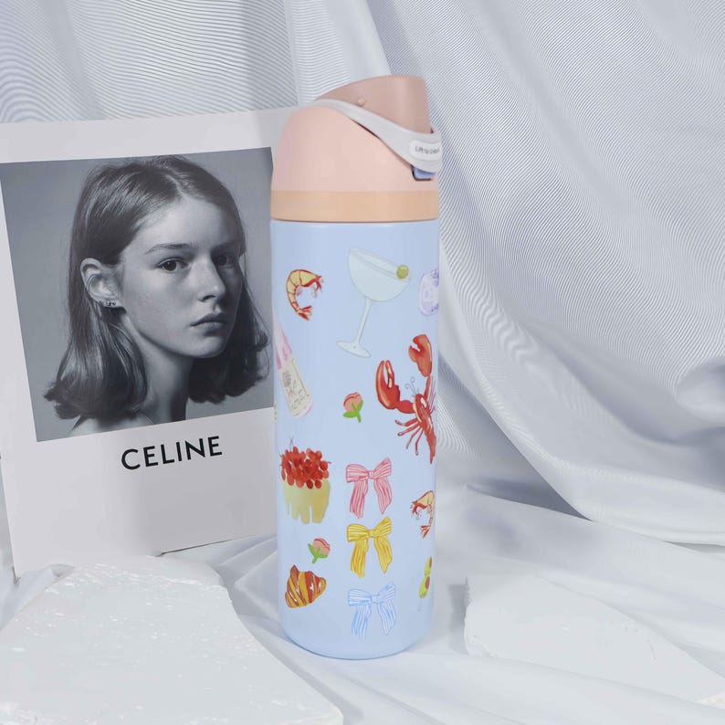 May include: A light blue insulated water bottle with a pink lid and a beige band. The bottle is decorated with illustrations of food and drinks, including a lobster, shrimp, and a martini glass. A black and white photo with the word "CELINE" is in the background.