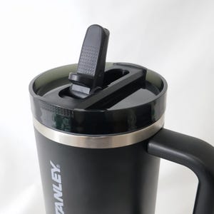 May include: Black insulated travel mug with a black handle and lid. The lid has a flip-top closure and a silver band around the top. The mug has the word "STANLEY" printed on the side.
