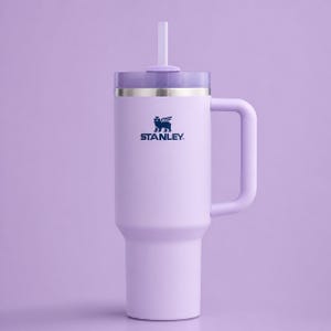 Puede incluir: Un vaso Stanley grande, de color lavanda, con asa y pajita. El vaso tiene tapa y una banda plateada cerca de la parte superior. El logotipo de Stanley est&aacute; impreso en azul en la parte delantera. El fondo es de color lavanda a juego.