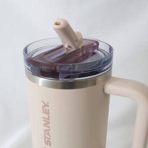 Pu&ograve; includere: Una tazza Stanley beige con manico e coperchio trasparente. Il coperchio ha un beccuccio ribaltabile e un interno viola scuro. Il logo Stanley &egrave; stampato verticalmente sul lato della tazza.