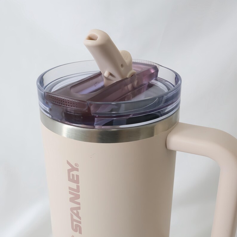 Pu&ograve; includere: Una tazza Stanley beige con manico e coperchio trasparente. Il coperchio ha un beccuccio ribaltabile e un interno viola scuro. Il logo Stanley &egrave; stampato verticalmente sul lato della tazza.