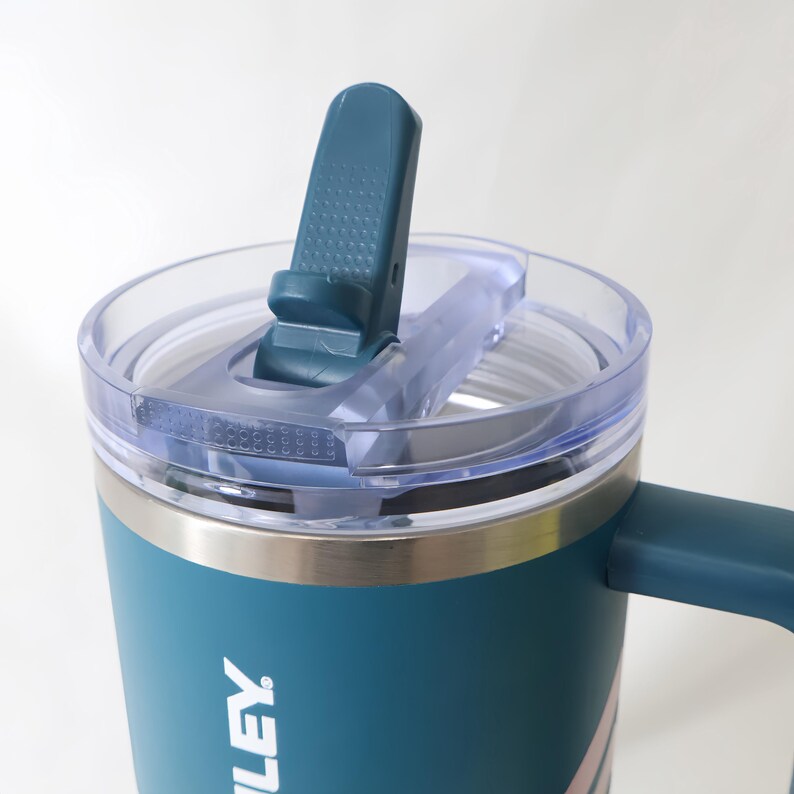 May include: A teal insulated travel mug with a clear lid and a teal flip-top closure. The mug has a silver band and a handle. The word "LEY" is visible on the side of the mug.
