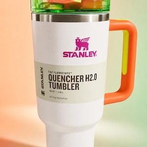 Pu&ograve; includere: Un bicchiere Stanley Quencher H2.0 bianco con un manico arancione brillante e un coperchio verde e rosa. Il bicchiere presenta il logo Stanley e il testo "QUENCHER H2.0 TUMBLER" e "1,18 L" stampati sul lato.