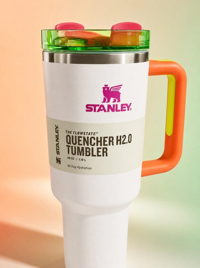Pu&ograve; includere: Un bicchiere Stanley Quencher H2.0 bianco con un manico arancione brillante e un coperchio verde e rosa. Il bicchiere presenta il logo Stanley e il testo "QUENCHER H2.0 TUMBLER" e "1,18 L" stampati sul lato.