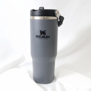 Op de afbeelding: Een grijze Stanley-beker met een zwart deksel en handvat. De beker heeft een zilveren rand en het Stanley-logo is zwart gedrukt. Ontworpen om dranken warm of koud te houden.