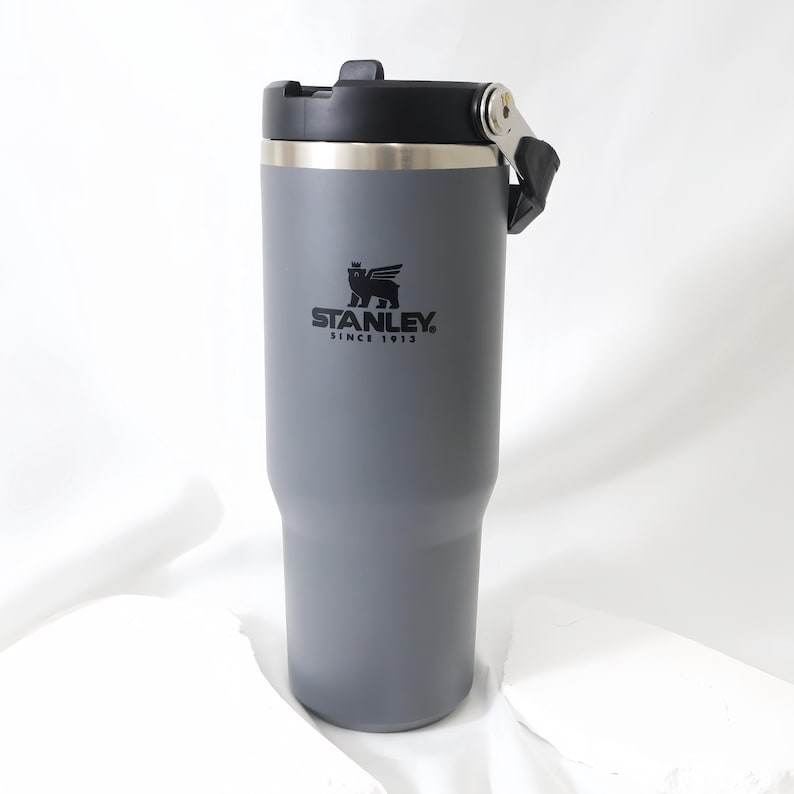 Op de afbeelding: Een grijze Stanley-beker met een zwart deksel en handvat. De beker heeft een zilveren rand en het Stanley-logo is zwart gedrukt. Ontworpen om dranken warm of koud te houden.