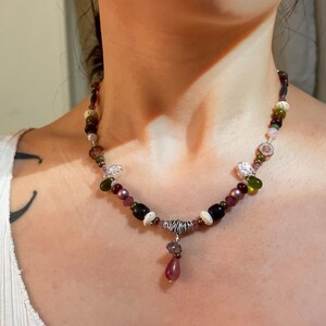 Puede incluir: Un collar de cuentas con una variedad de cuentas coloridas en tonos morado, verde, blanco y marrón. El collar tiene un colgante plateado con una cuenta en forma de lágrima. El collar se lleva alrededor del cuello.