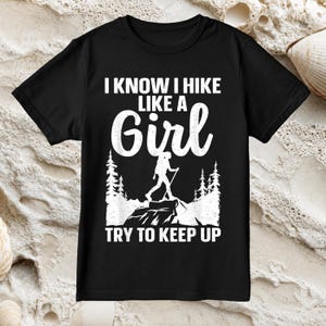 May include: A black short-sleeved t-shirt featuring white graphic text that reads I KNOW I HIKE LIKE A GIRL TRY TO KEEP UP. The design includes a white silhouette of a hiker with a backpack and trekking poles standing on a rock among pine trees.