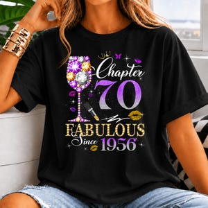 May include: A black t-shirt featuring a graphic design with a wine glass filled with purple and gold jewels, the text "Chapter 70", "FABULOUS", and "Since 1956". The design includes a lipstick, small butterflies, and lip prints in purple and gold.