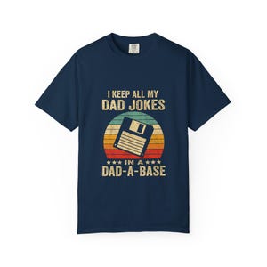 Op de afbeelding: Marineblauwe t-shirt met de tekst "I KEEP ALL MY DAD JOKES IN A DAD-A-BASE" en een retro afbeelding van een diskette over een zonsondergang. Het shirt heeft een ronde hals.
