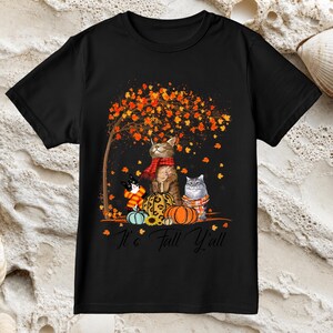 Puede incluir: Una camiseta negra de manga corta con un gráfico de tres gatos con bufandas sentados entre calabazas bajo un árbol de otoño con hojas naranjas. El texto It's Fall Y'all está impreso en una fuente cursiva negra en la parte inferior.
