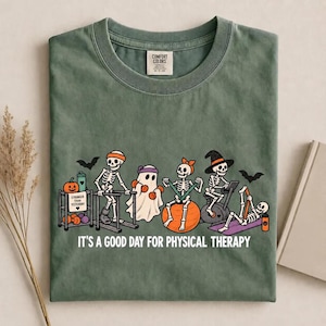 May include: A sage green crew neck t-shirt featuring a graphic of skeletons and a ghost performing physical therapy exercises. The text reads IT'S A GOOD DAY FOR PHYSICAL THERAPY. The design includes pumpkins, bats, and gym equipment in orange, black, and teal.
