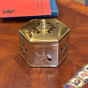 May include: A hexagonal brass box with a removable lid featuring a central knob. The sides and lid are decorated with cut-out floral patterns. The metallic surface has a polished finish, and the structure is composed of six flat panels.