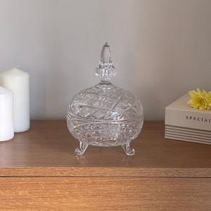 May include: A clear, round, footed glass candy dish with a removable lid featuring a pointed finial. The glass surface is decorated with a pressed geometric pattern of intersecting lines and starburst motifs, supported by three small, curved glass feet.