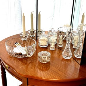 May include: A display of crystal glassware and decorative items on a wooden table. Includes a large bowl, small jars, vases, candle holders with candles, and a small round trinket box with a pearl necklace.
