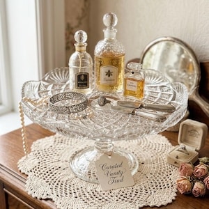Puede incluir: Un soporte para pasteles de vidrio prensado transparente con tres frascos de perfume de vidrio, una pulsera de filigrana plateada, un anillo y dos cuchillos de plata pequeños. Una etiqueta colgante dice Curated Vanity Find, junto a un collar de perlas.