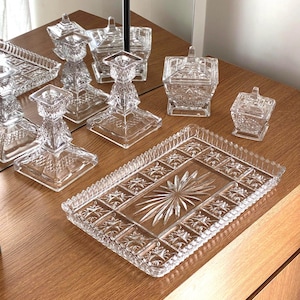 May include: A collection of clear pressed glass items including a rectangular tray with a central starburst pattern, two square candle holders with faceted stems, and two lidded square containers, all featuring intricate geometric relief designs.