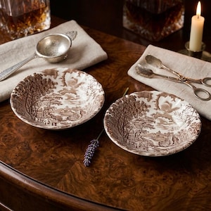 May include: Two round ceramic bowls featuring a brown and white transferware landscape pattern depicting trees, buildings, and a cloudy sky. The set includes a silver-toned tea strainer and a pair of silver-toned sugar tongs on linen napkins.
