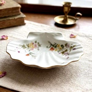 May include: A white porcelain dish shaped like a scallop shell with a gold-trimmed edge. The interior surface features a delicate floral pattern with small yellow, pink, and orange flowers and green leaves painted across the centre and sides.