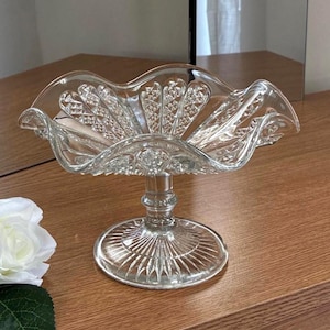 May include: A clear glass pedestal bowl featuring a ruffled, scalloped rim and a textured diamond pattern on the interior. The base is circular with a radiating sunburst design, connected to the bowl by a short, smooth, cylindrical glass stem.
