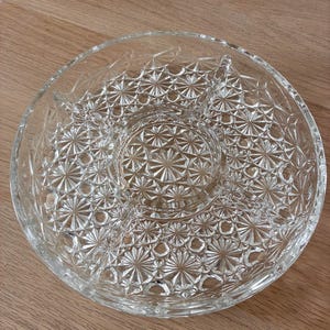 Könnte beinhalten: Eine runde Servierplatte aus klarem Glas mit unterteiltem Design, bestehend aus einem zentralen kreisförmigen Fach, das von drei äußeren Abschnitten umgeben ist. Die gesamte Oberfläche ist mit einem Stern- und Geometriemuster versehen.
