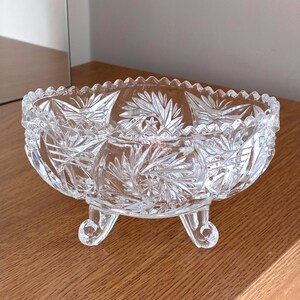 Könnte beinhalten: Eine klare, farblose Glasschale mit einem detaillierten, gepressten Sternen- und geometrischen Muster auf der Außenfläche. Die Schale steht auf drei kleinen, geschwungenen Füßen und hat einen gezackten, wellenförmigen Rand an der Oberseite.