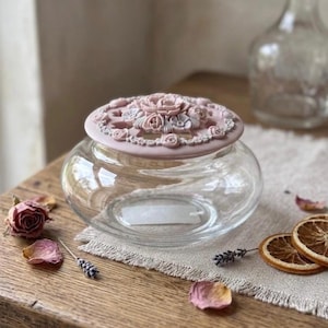 May include: A clear glass bowl featuring a removable, circular lid in a soft, dusty pink hue. The lid is adorned with intricate, raised floral carvings and decorative cutouts, providing a textured contrast to the smooth, transparent glass base.