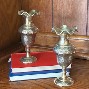 May include: Two matching brass vases with a metallic gold finish, featuring a flared, ruffled rim, a slender neck, and a bulbous body decorated with intricate floral engravings. Each vase stands on a circular pedestal base with leaf-like patterns.