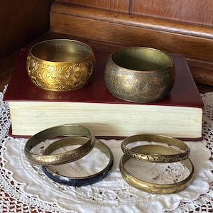 Op de afbeelding: Een collectie van zes metalen armbanden. Twee brede armbanden zijn voorzien van ingewikkelde bloem- en bladgravures. Vier dunnere armbanden bevatten variaties van gladde, getextureerde en patronen, waarvan er één een donkere afwerking heeft.
