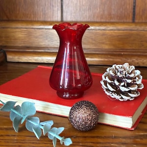May include: A small, translucent ruby red glass vase with a ruffled rim, positioned on a red hardback book alongside a brown glittery sphere, a pinecone with white-tipped scales, and a sprig of light green eucalyptus leaves.