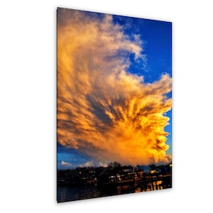 May include: A vertical wall art print featuring a dramatic sunset sky with large, golden-orange clouds contrasting against a deep blue background. Below the sky, a dark silhouette of a waterfront area with buildings and a pier is visible.