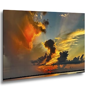 May include: A rectangular wall art print featuring a vibrant sunset sky with dramatic clouds in shades of deep orange, golden yellow, and dark gray. The horizon line is visible at the bottom, with light reflecting off the water surface below.