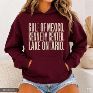 May include: A maroon hooded sweatshirt featuring a front kangaroo pocket and light beige text printed on the chest that reads: GULF OF MEXICO. KENNEDY CENTER. LAKE ONTARIO. The hoodie has long sleeves and a drawstring hood, constructed from fabric.