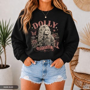 Op de afbeelding: Een zwart sweatshirt met lange mouwen en een grafische print van Dolly Parton met de tekst Dolly, 1946, 2026, Dolly Parton en Find out who you are and do it on purpose, omringd door illustraties van rozen, een vlinder en een cowboylaars.