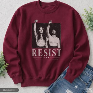 Op de afbeelding: Een bordeauxrode sweater met ronde hals en een zwart-wit afbeelding van twee personen met opgeheven vuisten. Onder de afbeelding staat het woord RESIST in witte hoofdletters, gevolgd door de woorden EQUALITY, JUSTICE en FREEDOM.