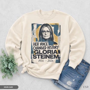 May include: A sand-colored crewneck sweatshirt featuring a graphic print of Gloria Steinem wearing glasses. The design includes blue and yellow abstract shapes, newspaper-style text, and the words "HER VOICE CHANGED HISTORY GLORIA STEINEM 1934-2026".