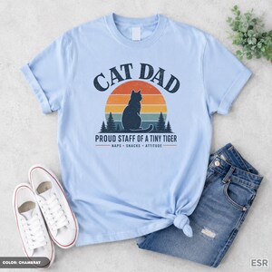 May include: A light blue t-shirt featuring a graphic of a cat silhouette in front of a sunset, with the text CAT DAD, PROUD STAFF OF A TINY TIGER, NAPS, SNACKS, ATTITUDE. The design includes pine trees and a vintage-style circular sunset background.