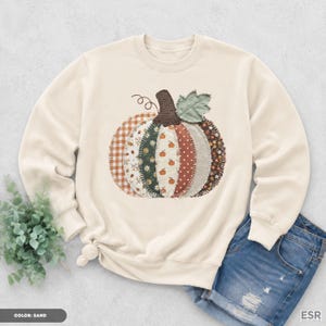 May include: A sand-coloured crewneck sweatshirt featuring a large, central graphic of a pumpkin composed of various fabric-style patches. The pumpkin design includes patterns like orange gingham, floral prints, polka dots, and stripes, topped with a dark brown stem and a green leaf.