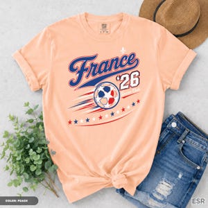 a peach-colored t-shirt with a graphic design of a soccer ball and stars, along with the text "France '26 Club" printed on it. The t-shirt is placed on a surface next to a pair of blue jeans and a straw hat.