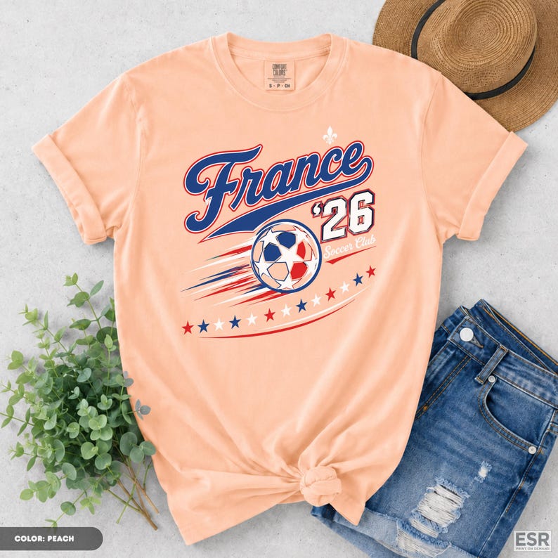 a peach-colored t-shirt with a graphic design of a soccer ball and stars, along with the text "France '26 Club" printed on it. The t-shirt is placed on a surface next to a pair of blue jeans and a straw hat.