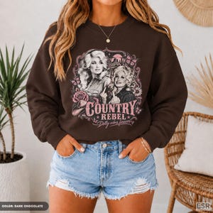 May include: A dark chocolate brown crewneck sweatshirt featuring a graphic print of two women with blonde hair, a cowboy hat, flowers, and butterflies. The text reads "COUNTRY REBEL" and "Dolly vibes forever" in a pink, decorative font style.