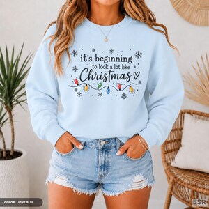 May include: A light blue crewneck sweatshirt featuring black text that reads "it's beginning to look a lot like Christmas" with a heart symbol and a graphic of colorful string lights and snowflakes printed across the center of the chest.