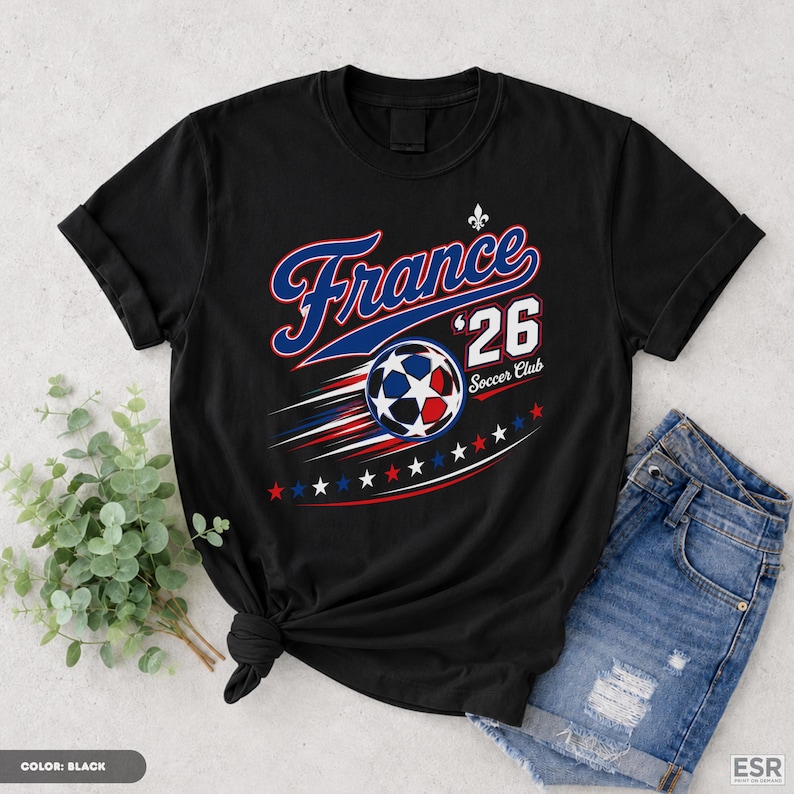 A black t-shirt with a graphic design featuring a soccer ball and the text "France '26 Soccer Club" is displayed on a white surface, along with a pair of denim shorts.