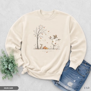 May include: A sand-colored crewneck sweatshirt featuring a central graphic illustration of a scarecrow, a rabbit, and pumpkins under a tree with falling leaves. The text "AUTUMN WHISPERS" is printed below the illustration in a simple font.