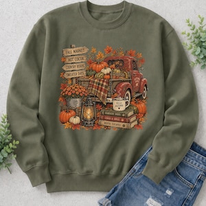 May include: An olive green crewneck sweatshirt featuring a graphic print of a red vintage truck loaded with pumpkins, a lantern, a stack of books, and a signpost with the text Fall Market, Hot Cocoa, Country Roads, and Sweater Days.