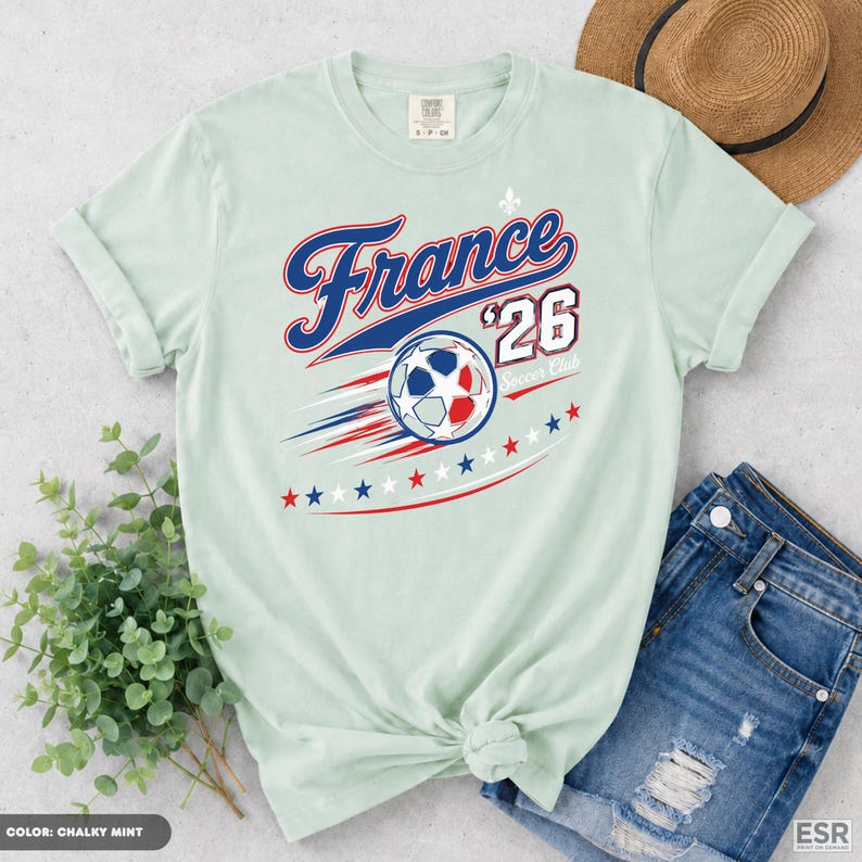 a light green t-shirt with a graphic design, a pair of blue jeans, and a straw hat, all laid out on a surface.